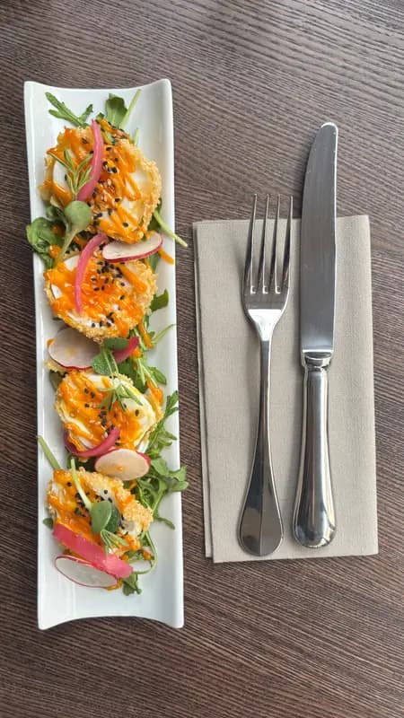 Crispy fried goat cheese rounds with sriracha drizzle, black sesame seeds, pickled radish, and arugula on elegant white rectangular plate.
