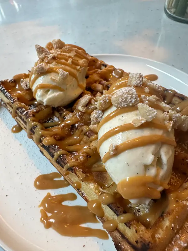 Waffle topped with two vanilla ice cream scoops, caramel drizzle, and crushed meringue pieces on a white plate.