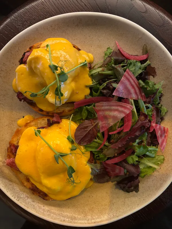 Eggs Benedict with hollandaise sauce, crispy bacon, and microgreens served with pickled beet salad on rustic ceramic plate.