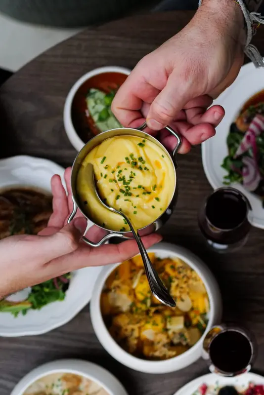 Creamy mashed potatoes garnished with chives in a mini cocotte, passed between diners at a restaurant table with various dishes.