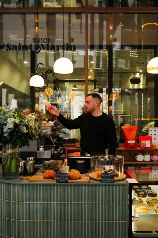 Barista preparing coffee at Maison Saint Martin café counter with croissants, brownies, and fresh flowers on display.