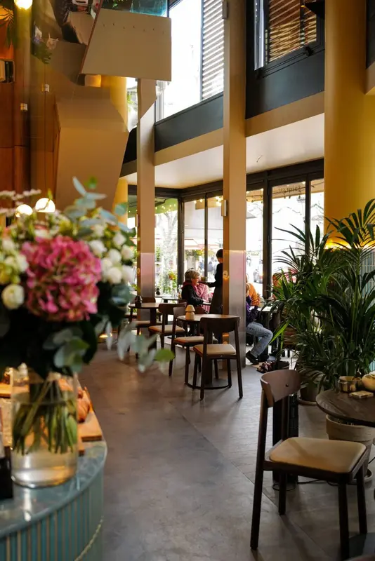 Bright restaurant interior with wood chairs, tropical plants, fresh flowers, and floor-to-ceiling windows overlooking a street.