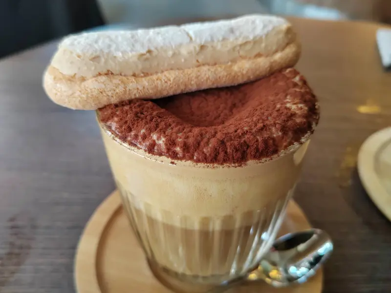 Tiramisu latte in a glass cup topped with cocoa powder and ladyfinger cookies, served on a wooden saucer