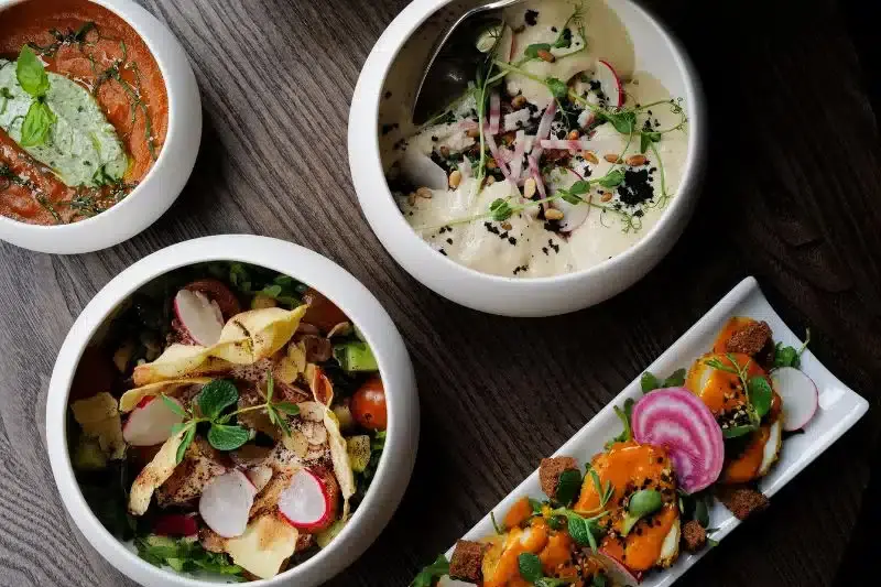 Four gourmet restaurant dishes including tomato soup, creamy hummus, fresh salad, and a colorful appetizer platter on a dark wood table.