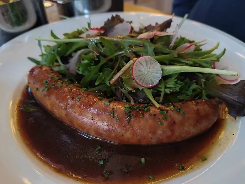 Pan-seared sausage in rich brown sauce topped with fresh arugula, radish salad, and chopped chives on white plate